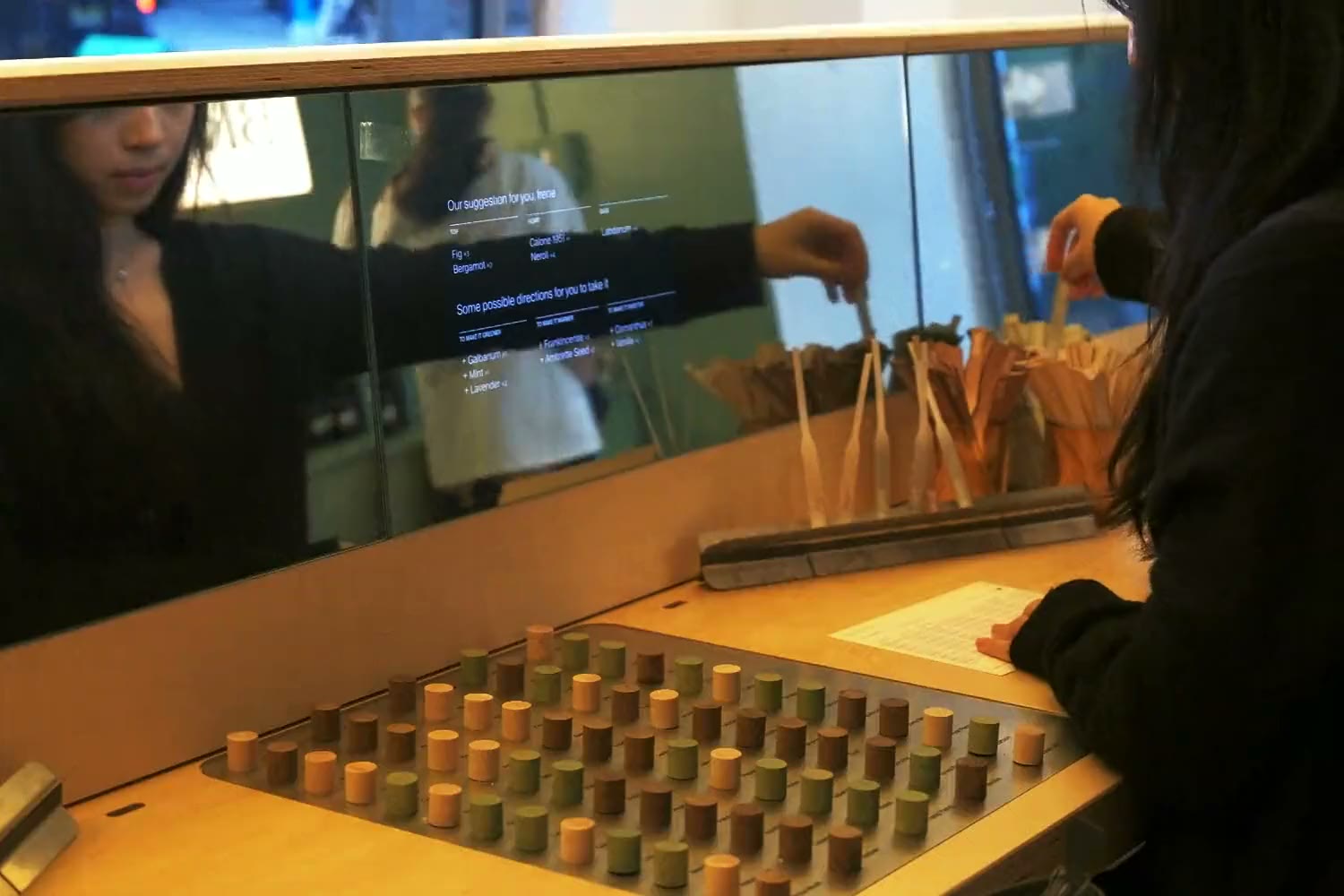 A guest choosing note samples from the tray set into the studio bench, then standing scent strips in the bench’s steel holder below the workstation screen’s suggested notes